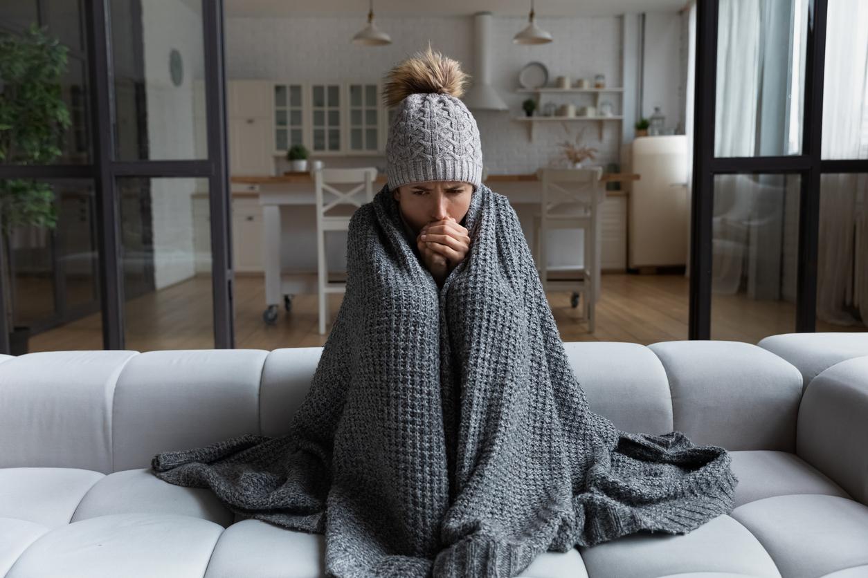 A young woman, dressed in winter clothes, freezing on the couch wrapped in a blanket with a broken heating system