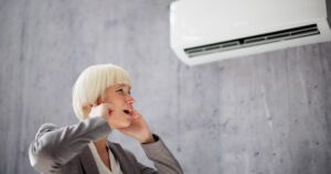 Woman covering her ears beneath a noisy wall-mounted air conditioner.